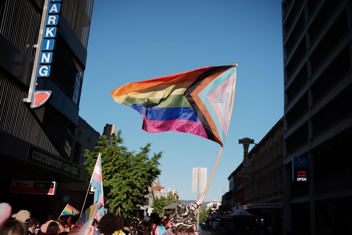 Progress Pride flag held high against blue sky at a street march, with trans pride flag visible in the background.